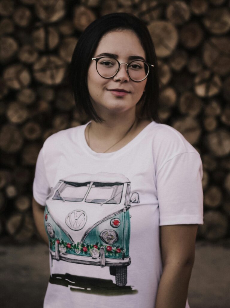 Smiling woman in glasses and VW T-shirt posing against a log backdrop.